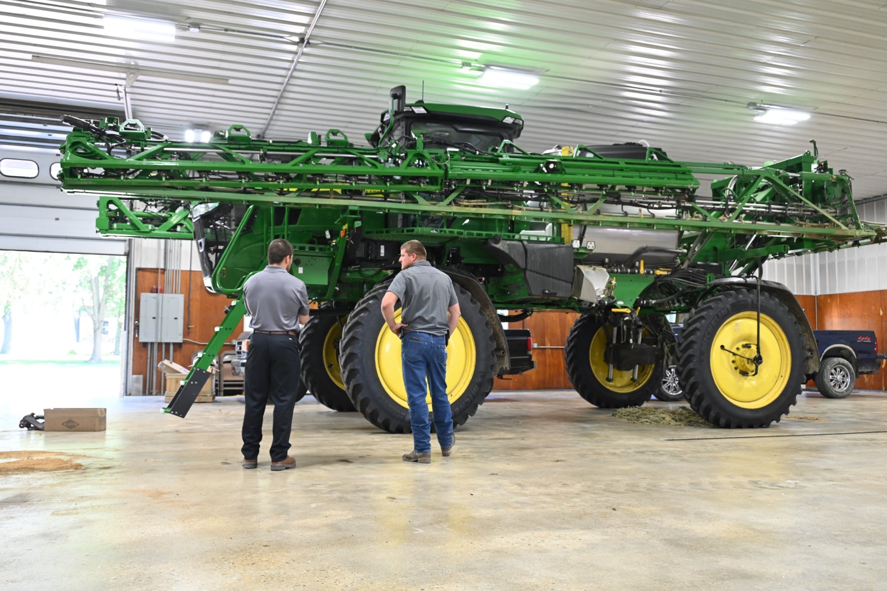 Two men inspect a large green John Deere sprayer with yellow wheels in a service bay.