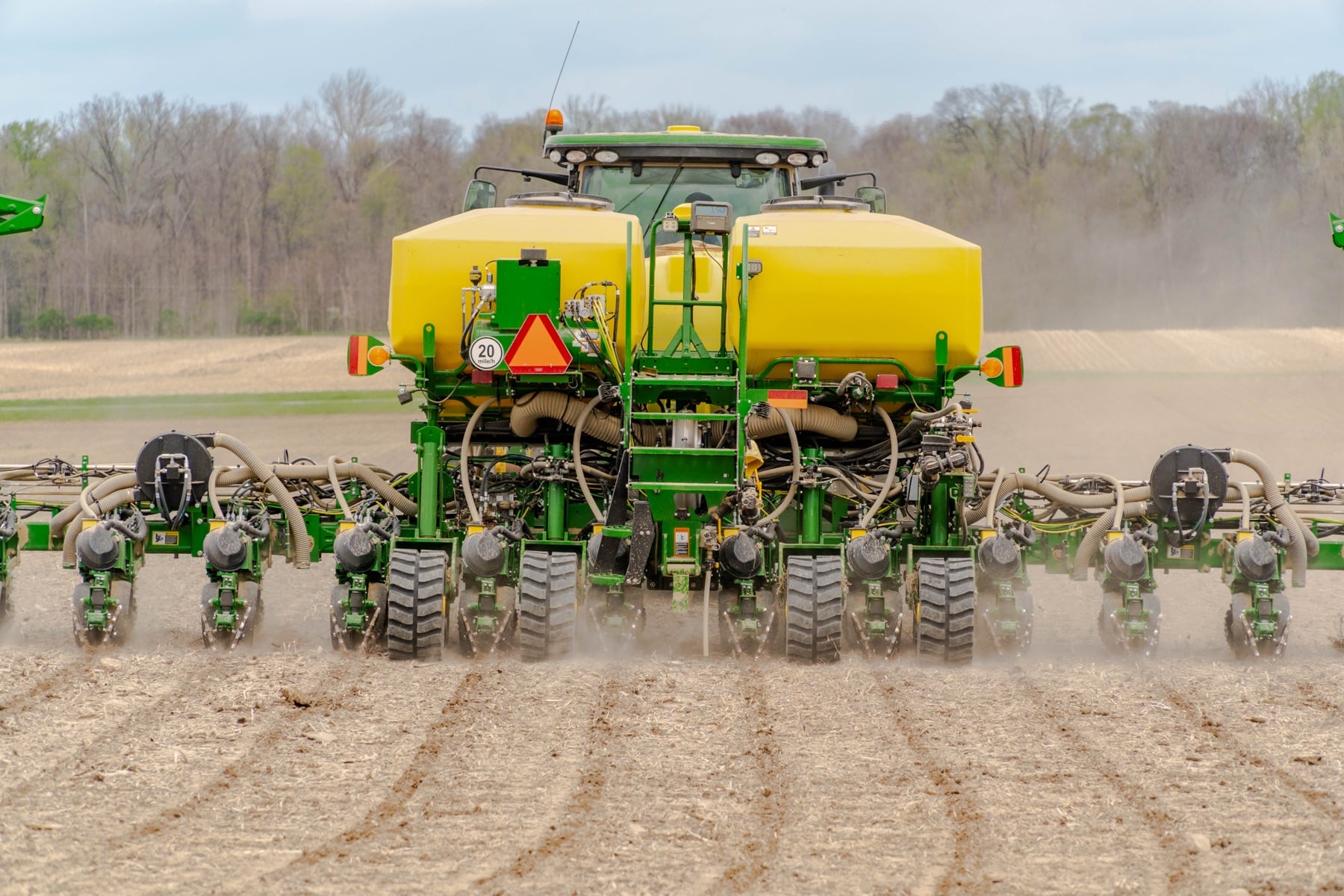 A green and yellow John Deere tractor pulls a large planter with multiple row units across a tilled field, kicking up dust.