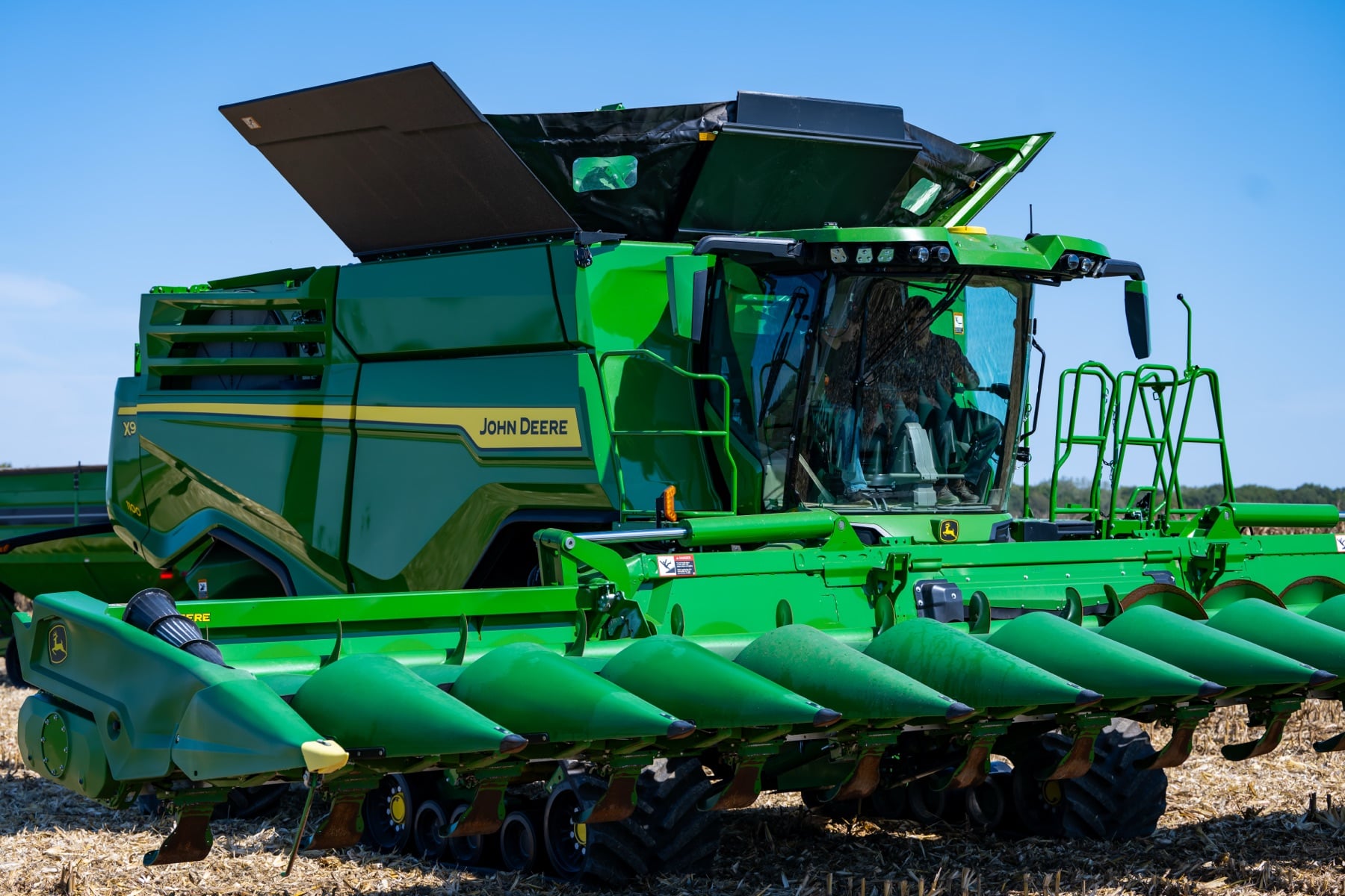 A green John Deere X9 combine with a corn head sits in a harvested field under a clear blue sky.