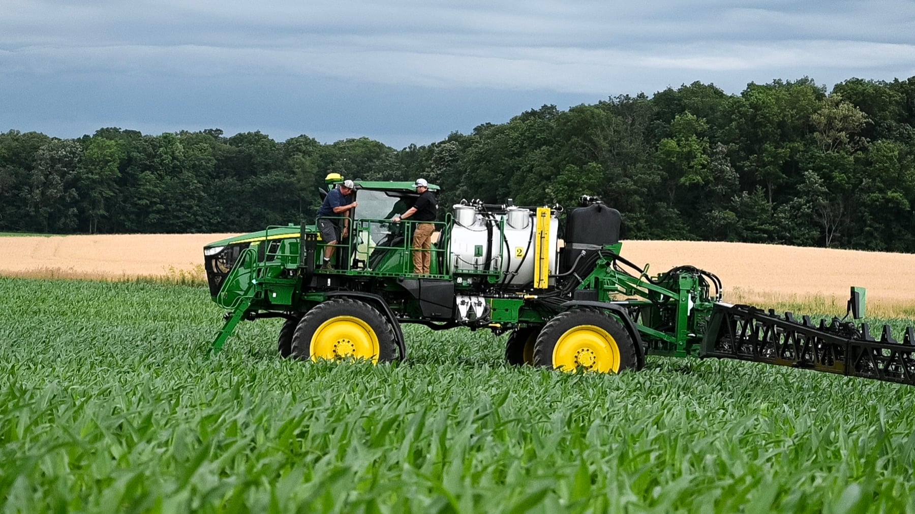 Two men stand on a green John Deere self-propelled sprayer in a field of green crops, with a golden field and trees behind.