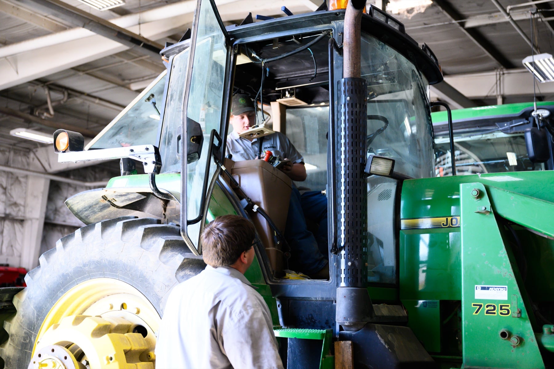 Two technicians inspect a large green John Deere agricultural tractor in a service bay.