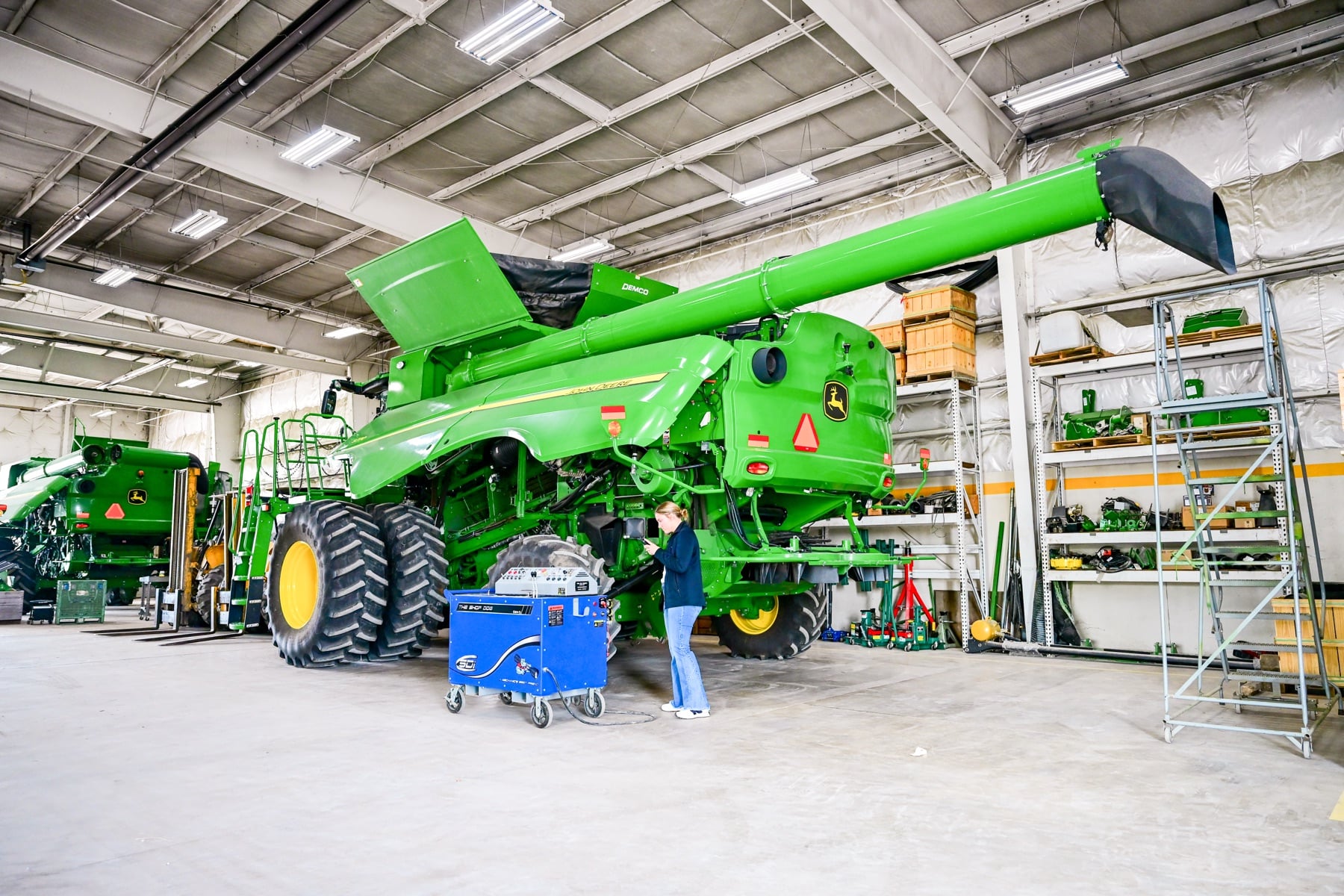 A female technician inspects a large green John Deere combine harvester in a service bay, using diagnostic equipment.