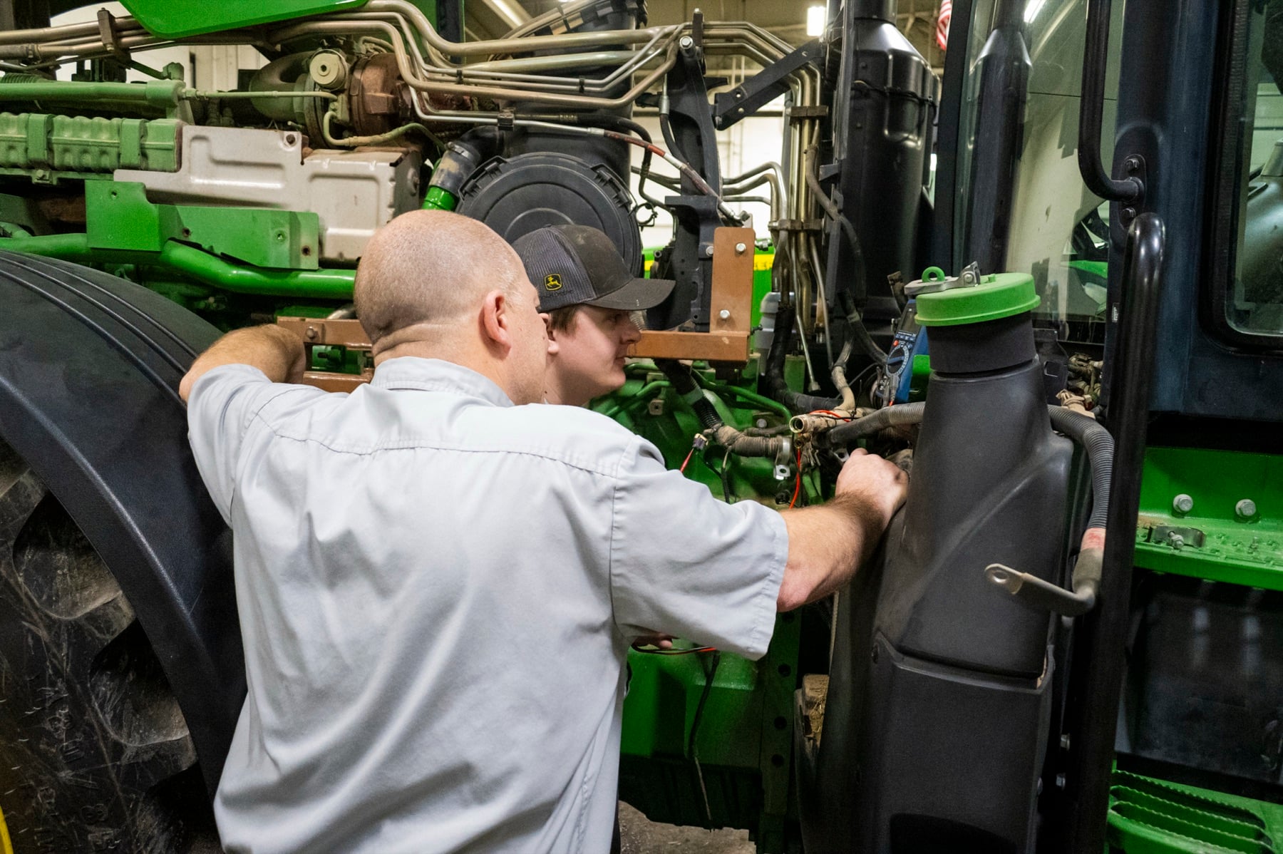 Two technicians inspect the engine of a large green John Deere tractor in a workshop.