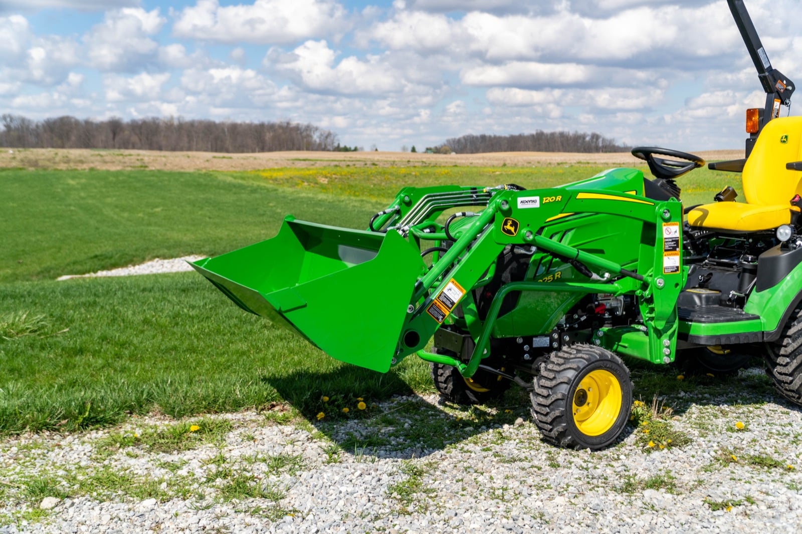 John Deere 1025R with a 120R loader on a gravel lot by an open field