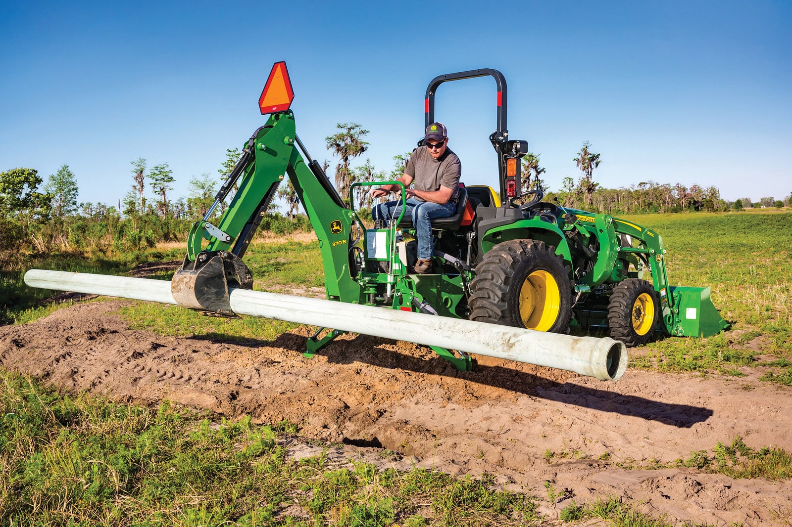 A John Deere 3038E with a backhoe carrying pipe